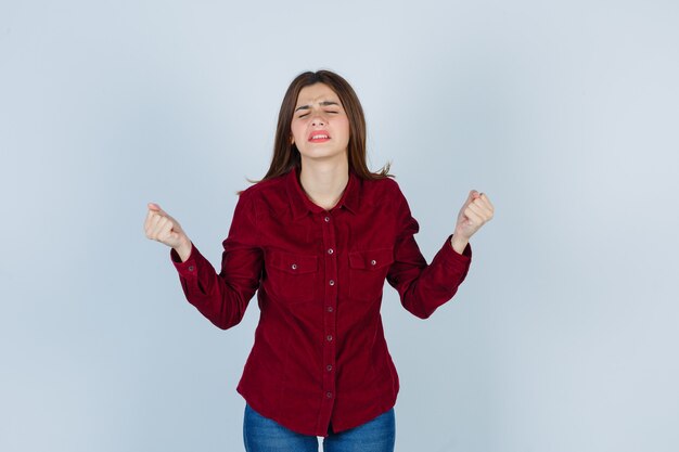 chica manteniendo los puños cerrados, cerrando los ojos con una blusa color burdeos y luciendo frustrada.