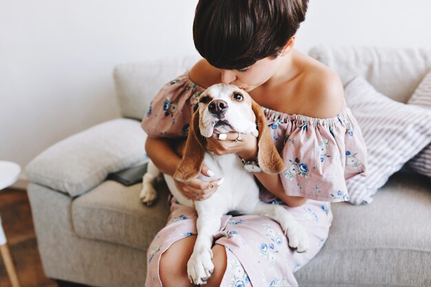 Chica maravillosa con peinado corto de moda besando a perro beagle mientras está sentado en el sofá gris