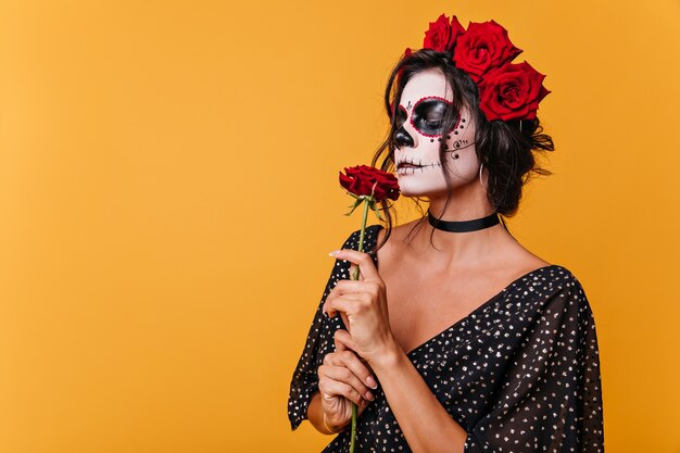 Chica con máscara de zombie para Halloween huele a rosa fragante. Modelo en vestido negro sobre pared naranja