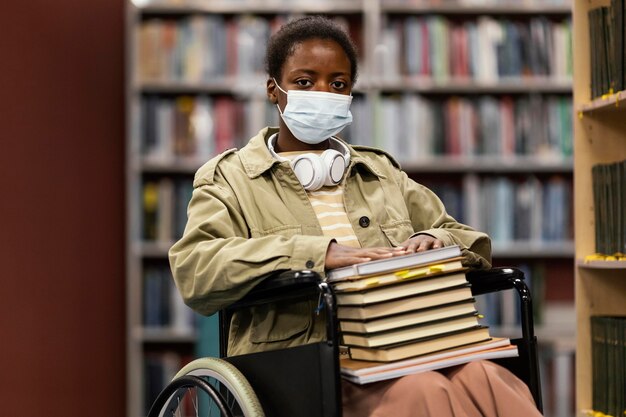 Chica con mascarilla en silla de ruedas sosteniendo un montón de libros