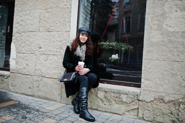 Chica mexicana rizada con gorra de cuero y taza de café de plástico a mano caminando por las calles de la ciudad