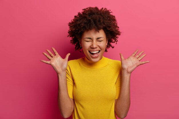 La chica milenaria alegre y optimista se ríe y levanta las palmas, tiene una expresión de cara muy feliz, mira una escena divertida, vestida con una camiseta amarilla, aislada en una pared rosa. Concepto de emociones positivas