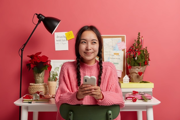 Foto gratuita una chica milenaria alegre y soñadora se sienta relajada contra el escritorio, charla con amigos, tiene un descanso para enviar mensajes, decora el lugar de trabajo para navidad