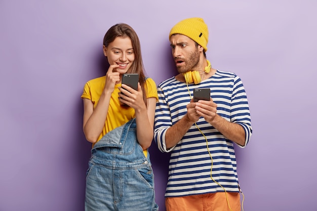 La chica milenaria mira positivamente el dispositivo del teléfono inteligente, un tipo sorprendido y desconcertado con celular, de pie cerca uno del otro contra la pared púrpura. Juventud con tecnologías modernas. Pareja adicta