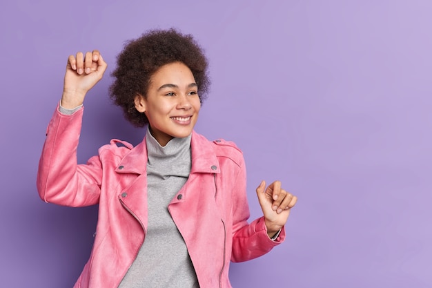 Chica milenaria positiva con cabello rizado levanta los brazos y baila sin preocupaciones tiene un estado de ánimo optimista y tontos vestidos con una chaqueta rosa