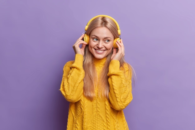 Chica milenaria positiva disfruta de música agradable a través de auriculares, está de buen humor, sonríe felizmente vestida con un jersey amarillo equipado