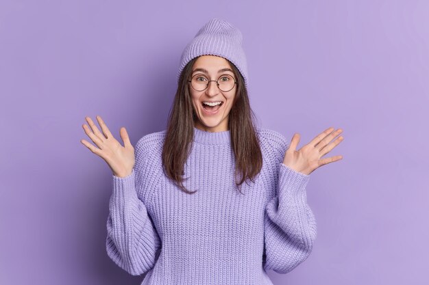 La chica millennial morena positiva levanta las palmas de las manos y reacciona ante algo impresionante y se pone muy feliz con sombrero y suéter.