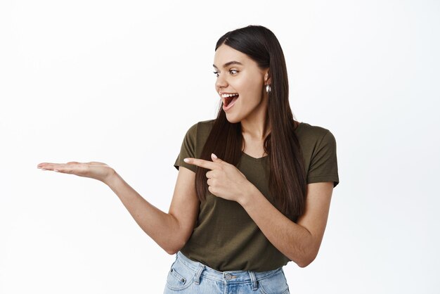 La chica mira emocionada su palma con el anuncio del artículo apuntando a la mano contra el espacio de la copia de pie sobre fondo blanco