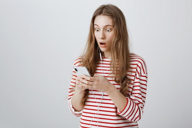 Chica mira fijamente el teléfono móvil sorprendida, leyendo noticias impactantes