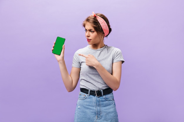 Chica mira con malentendido en smartphone. Mujer joven moderna en camiseta gris y falda de mezclilla con cinturón ancho posando.