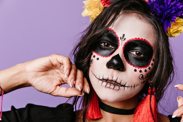 Chica con mirada mágica posa en la pared púrpura. Mujer con flores en el pelo vestida para Halloween.