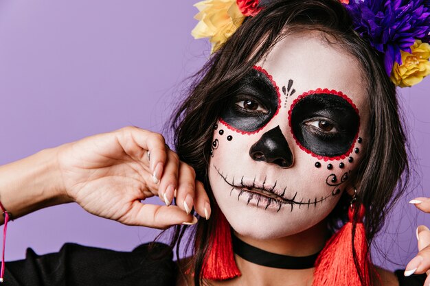 Chica con mirada mágica posa en la pared púrpura. Mujer con flores en el pelo vestida para Halloween.