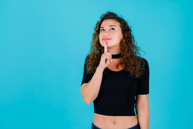 Chica mirando hacia arriba está poniendo el dedo índice en los labios sobre fondo azul.