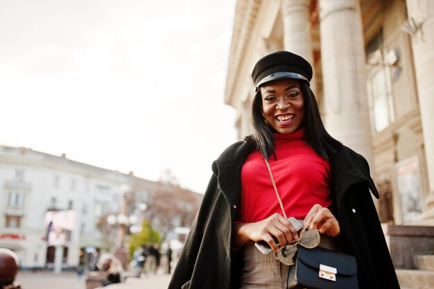 Chica de moda afroamericana con abrigo y gorra de repartidor de periódicos posada en la calle