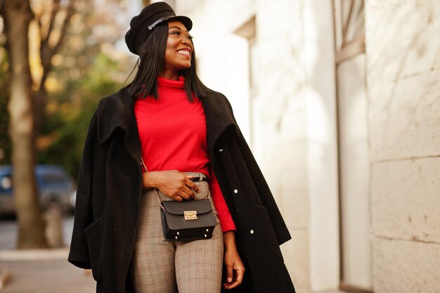 Chica de moda afroamericana con abrigo y gorra de repartidor de periódicos posada en la calle