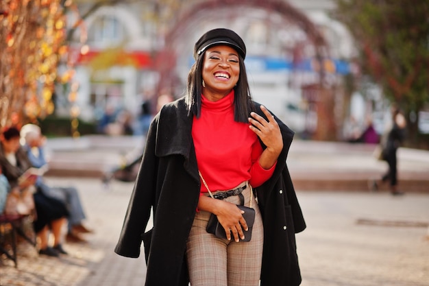 Chica de moda afroamericana con abrigo y gorra de vendedor de periódicos con bolso posado en la calle