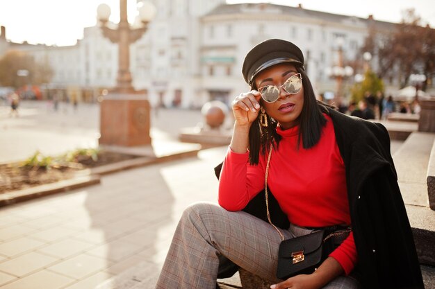 Chica de moda afroamericana en abrigo y gorra de vendedor de periódicos gafas de sol posadas en la calle