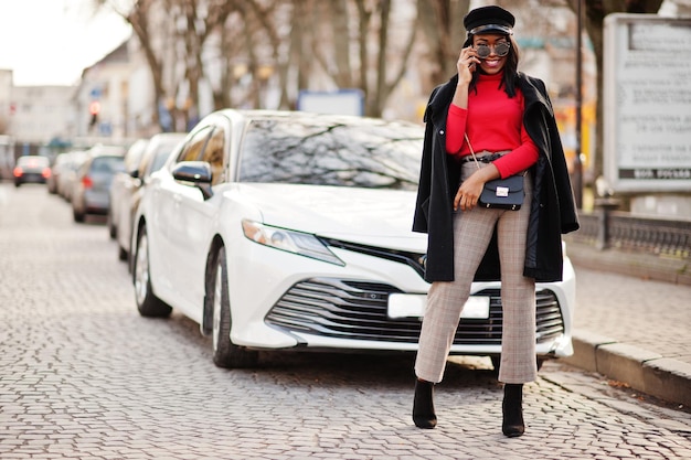 Chica de moda afroamericana con gorra de vendedor de periódicos y gafas de sol posadas en la calle contra un coche de negocios blanco