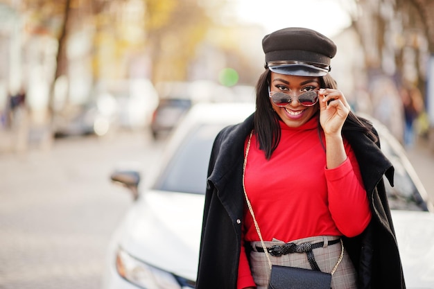 Chica de moda afroamericana con gorra de vendedor de periódicos y gafas de sol posadas en la calle contra un coche de negocios blanco