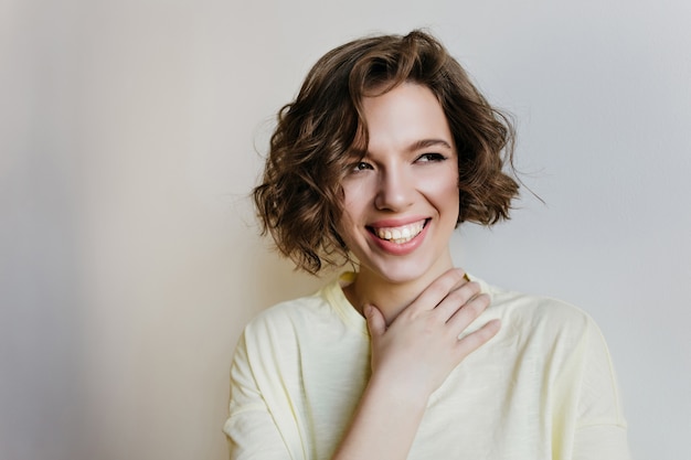 Chica de moda con cabello oscuro brillante mirando a otro lado con sonrisa feliz. Foto interior de mujer blanca entusiasta aislada en pared ligera.