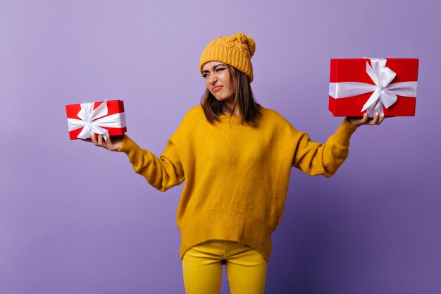 Chica de moda elegante en ropa amarilla con regalos y haciendo muecas. Retrato de interior de hermosa mujer morena en traje casual.