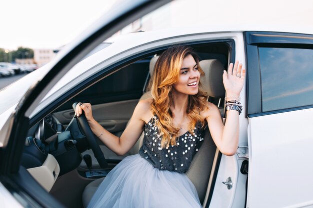 Chica de moda en falda de tul conduciendo coche blanco. Ella gratifica a alguien a su lado.