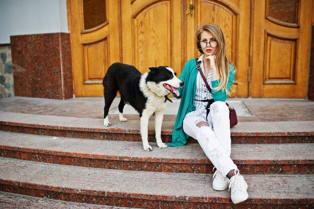 Chica de moda con gafas y jeans rotos con perro husky laika ruso con una correa contra la puerta de madera en la calle de la ciudad Amigo humano con tema animal