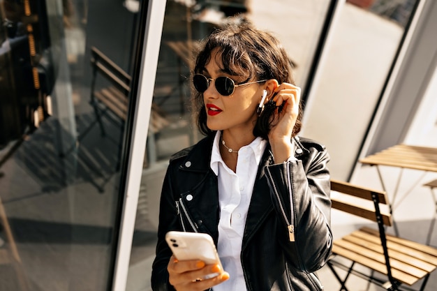 Chica de moda con peinado corto y labios rojos con chaqueta de cuero y gafas negras escuchando música y usando un teléfono inteligente en la luz del sol en el fondo de la ciudad