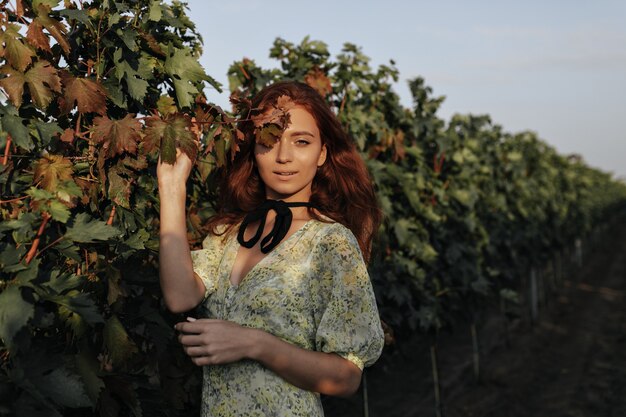 Chica de moda con peinado largo rojo y vendaje negro en el cuello con ropa verde de moda ligera mirando al frente en viñedos