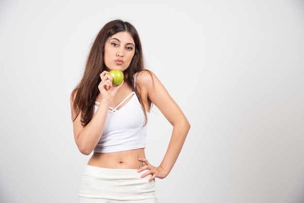 Chica de moda en ropa deportiva sosteniendo una manzana en la pared gris.