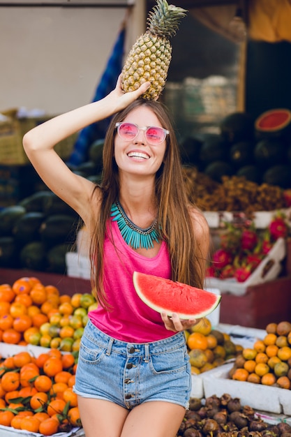 Chica de moda de verano disfrutando en el mercado de frutas tropicales. Ella sostiene ananas en la cabeza y una rodaja de sandía en la mano detrás