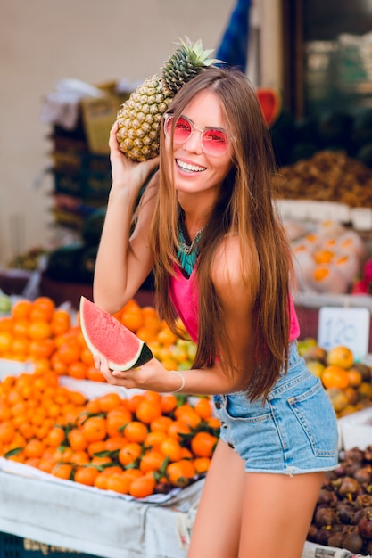 Chica de moda de verano está posando con ananas y una rodaja de sandía en el mercado de frutas tropicales