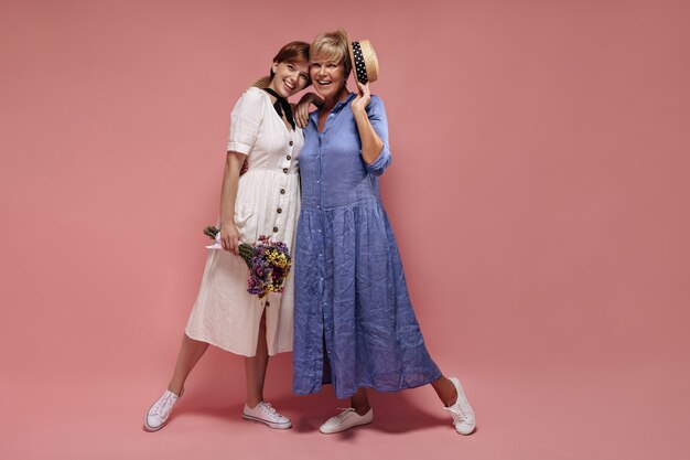 Chica de moda en vestido blanco y zapatillas de deporte con flores silvestres y sonriendo con una dama rubia en traje azul y sombrero de paja sobre fondo rosa.