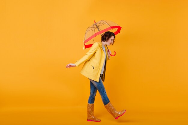 Chica de moda en zapatos de goma y chaqueta amarilla mirando hacia abajo mientras posa con paraguas. Foto de estudio de mujer de pelo corto rizado en jeans caminando con sombrilla.