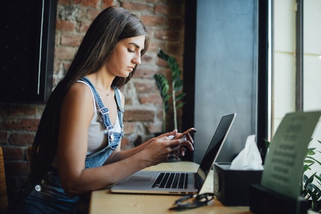 Chica modelo joven cansada está sentada en el café frente a la ventana trabaja en su computadora portátil y toma una bebida fresca