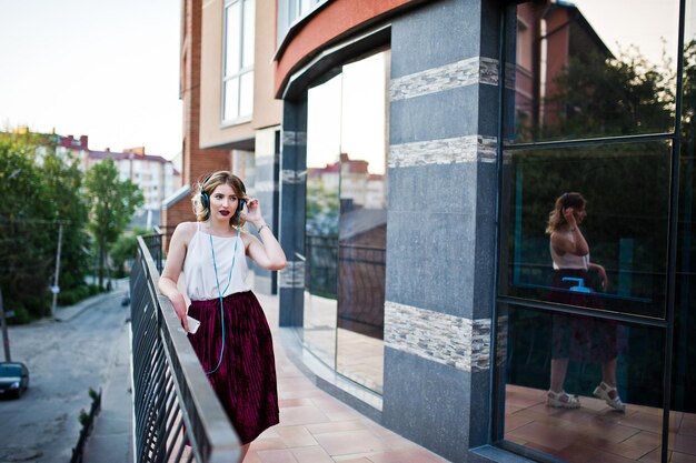 Chica modelo rubia de moda y hermosa en elegante blusa blanca de falda de terciopelo rojo posando con teléfono y auriculares contra las ventanas del edificio de la ciudad