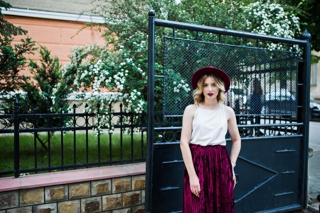 Chica modelo rubia de moda y hermosa en elegante falda de terciopelo rojo blusa blanca y sombrero posando al aire libre contra las puertas