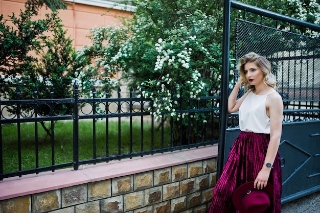 Chica modelo rubia de moda y hermosa en elegante falda de terciopelo rojo blusa blanca y sombrero posando al aire libre contra las puertas