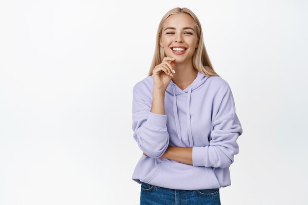 Chica moderna y elegante con corte de pelo largo rubio, sonriendo, mirando confiado, de pie en blanco