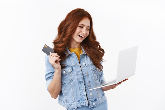 Chica moderna y elegante elige un nuevo atuendo en la tienda de Internet, compra en línea, sostiene la computadora portátil y agita la tarjeta de crédito negra con una sonrisa encantada y satisfecha, ingresa el número de cuenta bancaria, mira la pantalla del cuaderno