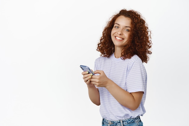 Chica moderna sonriente usando teléfono móvil, chateando y luciendo feliz, concepto de tecnología celular, usando camiseta.