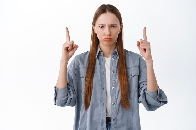 Chica molesta o enojada con el ceño fruncido, enfurruñada y señalando con el dedo algo malo, estar celosa, querer algo, quejarse, de pie decepcionada contra la pared blanca