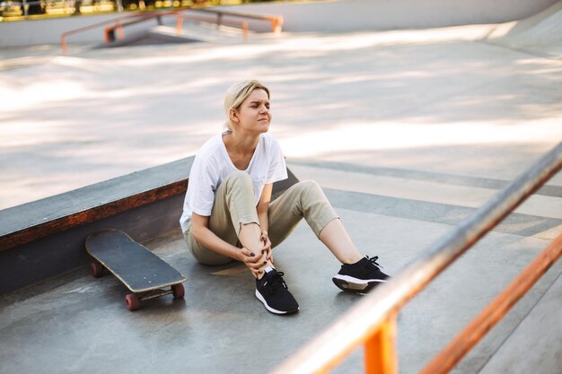 Chica molesta tristemente cerrando los ojos sosteniendo su pierna dolorosa con patineta cerca del skatepark aislado