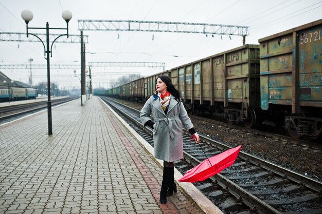 Chica morena en abrigo gris con paraguas rojo en la estación de tren