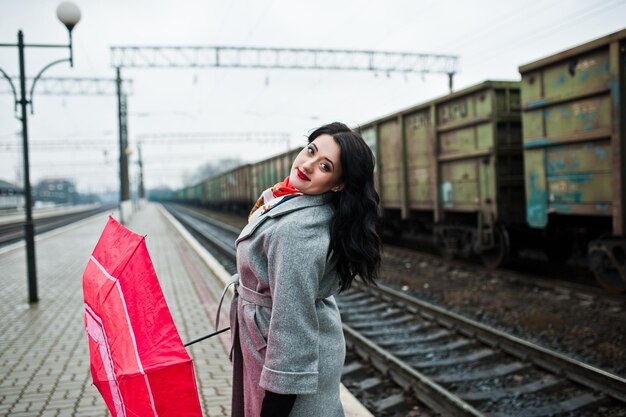 Chica morena en abrigo gris con paraguas rojo en la estación de tren