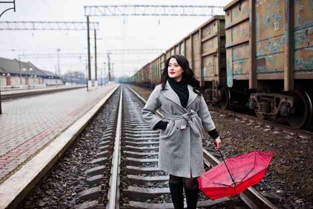 Chica morena en abrigo gris con paraguas rojo en la estación de tren