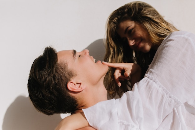 Chica morena alegre en blusa vintage de moda divirtiéndose con su novio tocando juguetonamente su barbilla. Retrato de pareja amorosa abrazándose y sonriendo delante de la pared blanca.