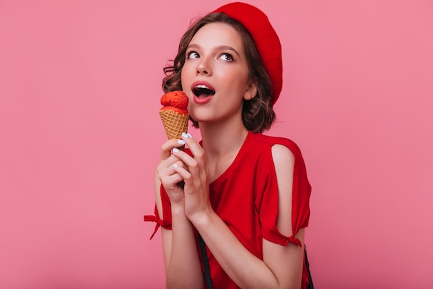 Chica morena alegre en camiseta roja posando con helado. Foto interior de una dama encantadora con boina comiendo postre.