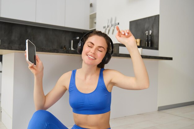 Chica morena alegre hace ejercicio en casa usa auriculares inalámbricos y baila sostiene un teléfono inteligente