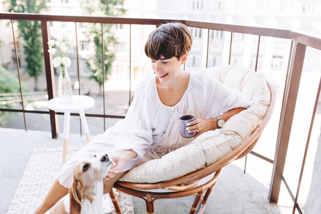 Chica morena alegre sosteniendo una taza de café acariciando el perrito divertido que mira hacia arriba con placer